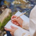Frau mit blonden Haaren sitzt an felsigem Ufer und schreibt in ein Notizbuch