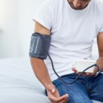 Junger Mann sitzt auf dem Bett und misst mit einem Oberarm-Messgerät seinen Blutdruck