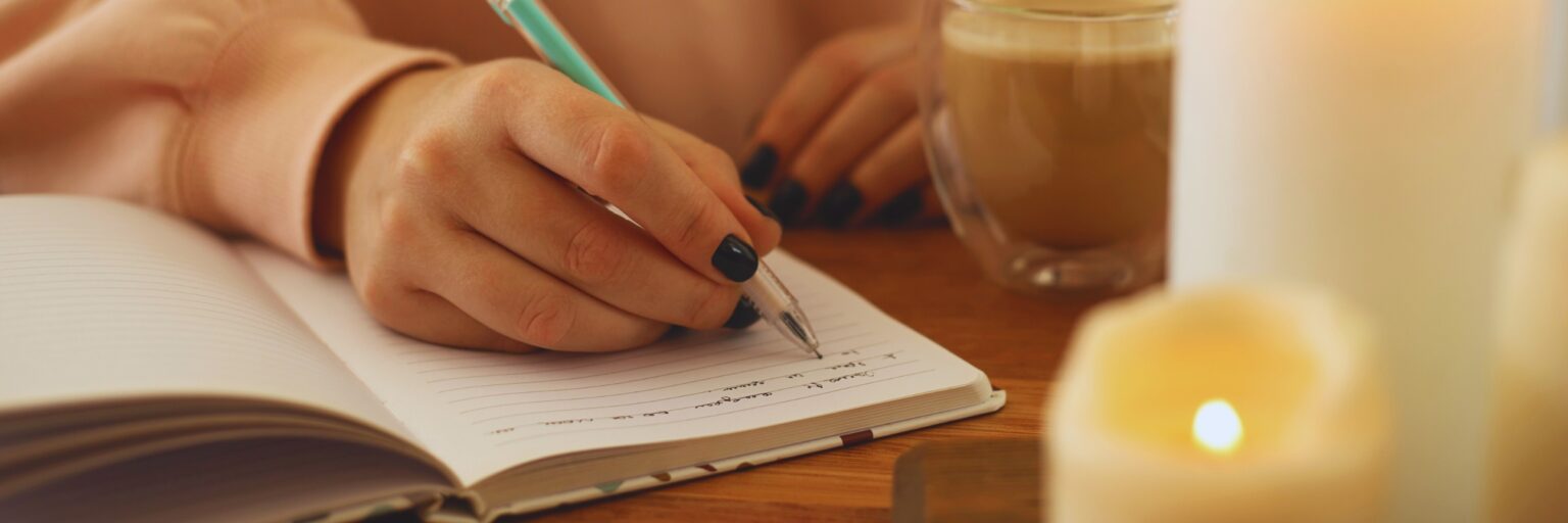 Hand schreibt in ein Notizbuch, daneben Kaffee und eine Kerze