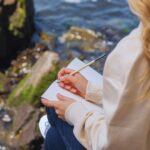 Frau mit blonden Haaren sitzt an felsigem Ufer und schreibt in ein Notizbuch