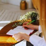 Frau mit langen roten Haaren schreibt an einem runden Holztisch in ein Notizbuch