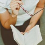 Frau sitzt auf dem Boden, hält ein aufgeschlagenes Buch und eine Brille in der Hand