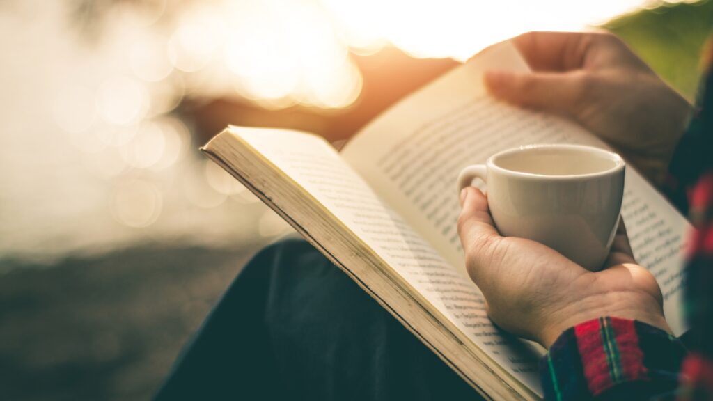 Person liest draußen ein Buch und hält eine Tasse in der Hand