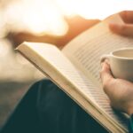 Person liest draußen ein Buch und hält eine Tasse in der Hand