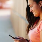 Lächelnde junge Frau im rosa Sportshirt schaut draußen auf ihr Smartphone