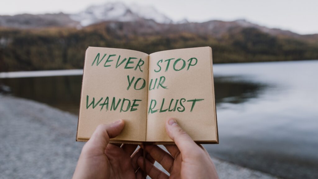 Hände halten ein Notizbuch mit der Aufschrift „Never stop your wanderlust“ vor einem Bergsee