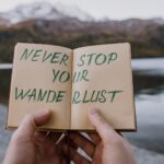 Hände halten ein Notizbuch mit der Aufschrift „Never stop your wanderlust“ vor einem Bergsee