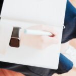 Person sitzt auf einem Holzboden und schreibt mit einem Stift in ein helles Notizbuch
