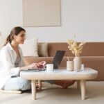Lächelnde Frau sitzt auf dem Boden im hellen Wohnzimmer und arbeitet am Laptop