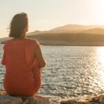 Frau im orangefarbenen Kleid sitzt auf Felsen und blickt bei Sonnenuntergang aufs Meer